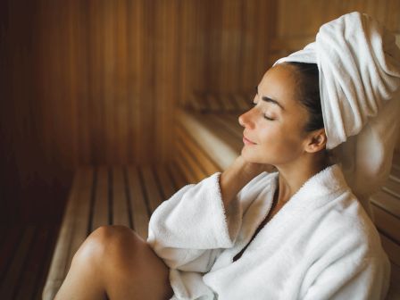 A woman in a spa, wrapped in a white towel, sits with eyes closed and relaxes in warm, cozy light.