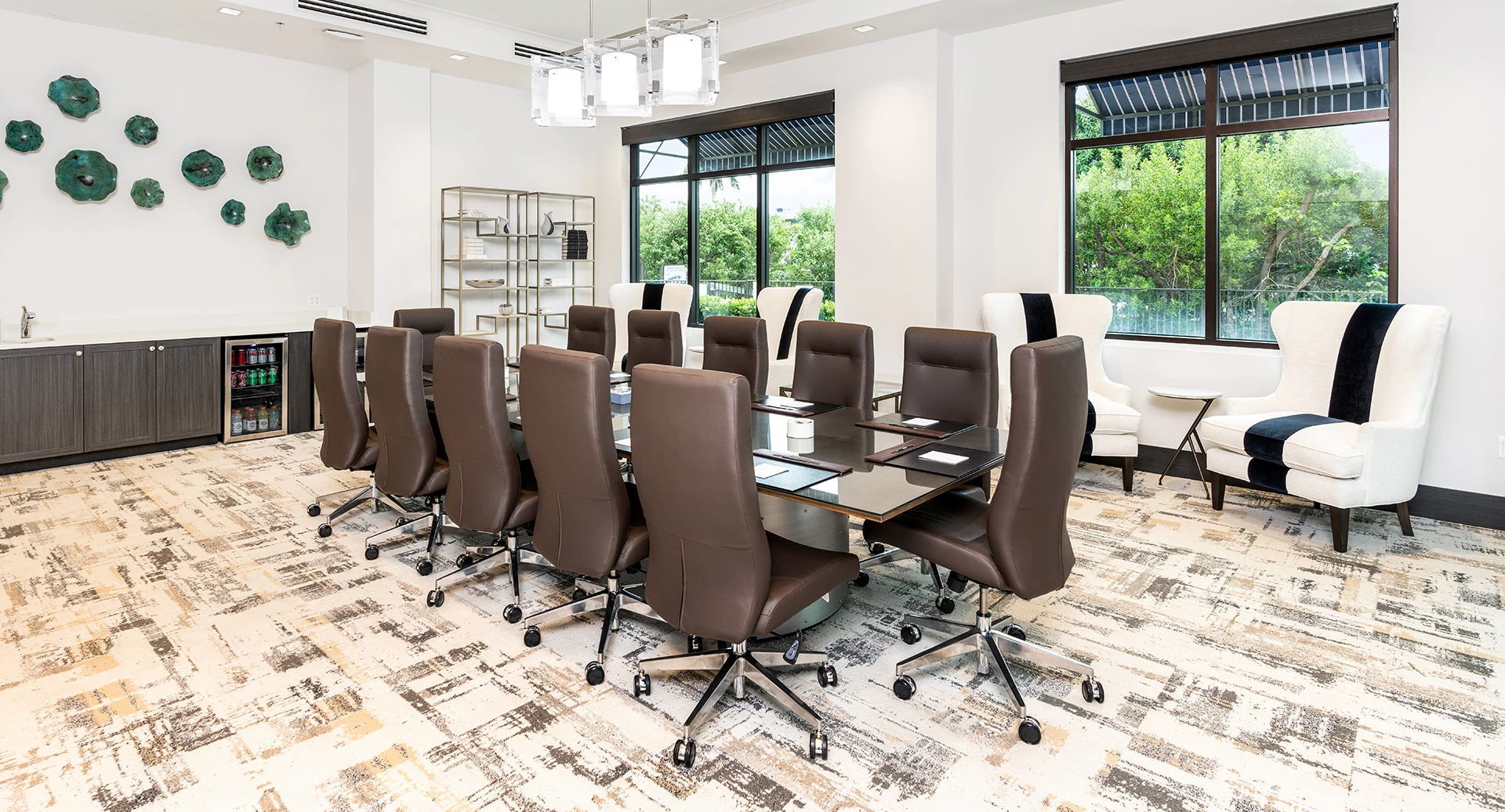 A modern conference room with a long dark wood table, brown rolling chairs, a sideboard, large windows, and white accent walls.