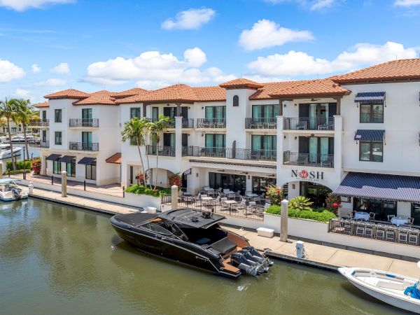 A sunny waterfront scene with a white Mediterranean-style building, palm trees, patios, and a docked black speedboat along a canal.