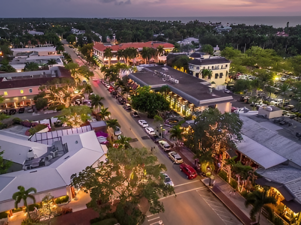 A bustling coastal town at dusk, palm-lined streets, pastel buildings, and warm storefront lights glow along a grid of calm avenues.