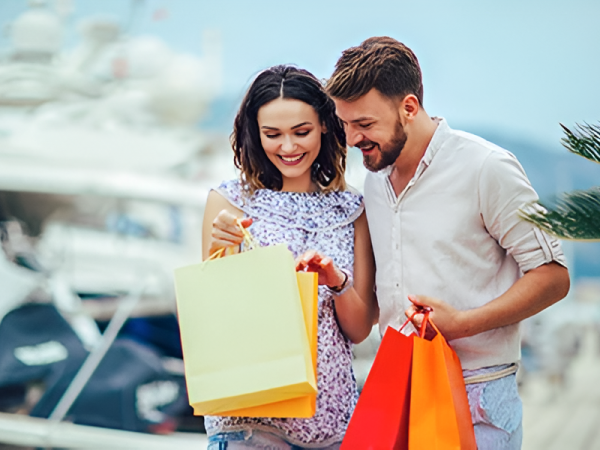 A smiling couple carrying colorful shopping bags near a marina with boats; they seem delighted by their purchases.