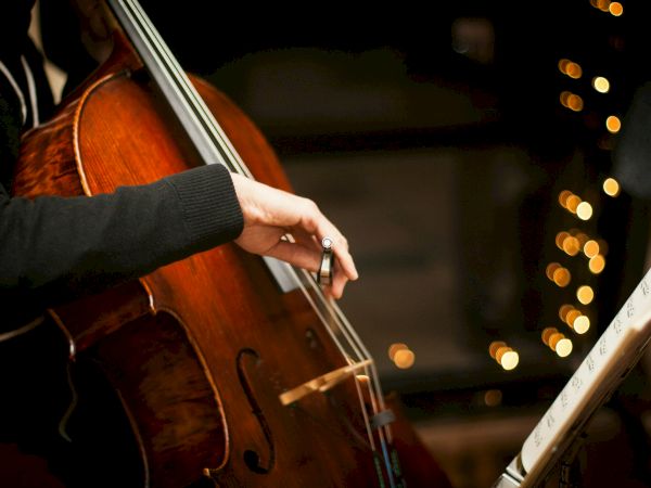 A close-up of a cellist's hands bowing a cello, with sheet music and warm bokeh lights in the background.