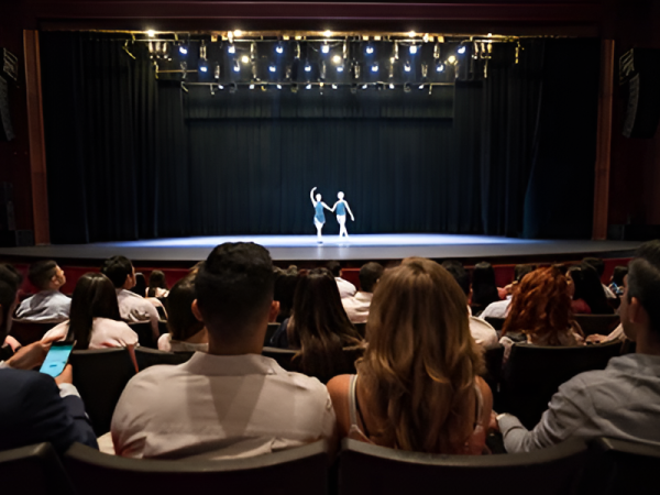 A couple dances center stage under bright stage lights while an audience watches from rows in a theater, evening recital vibe.