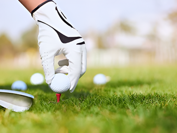 A gloved hand places a golf ball on a tee, ready for a short drive, with a club face approaching on a grassy practice area.