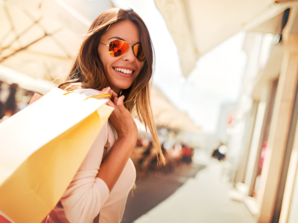 A smiling woman in sunglasses walks an outdoor shopping street with yellow shopping bags over her shoulder, sunny and vibrant.