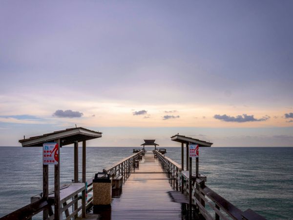 A wooden pier extends into calm sea at sunset, with small shelters on each side and a hazy, colorful sky above.