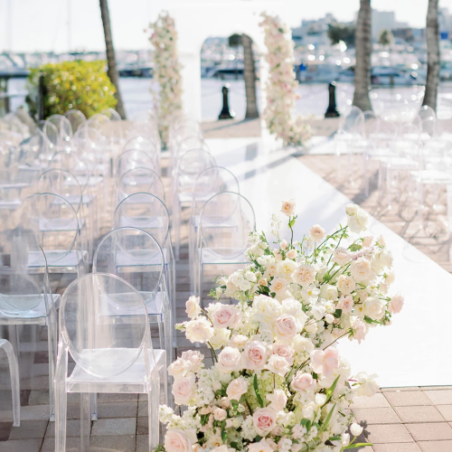 Elegant outdoor wedding setup by the harbor with clear acrylic chairs and a white aisle lined with pale pink roses and greenery.
