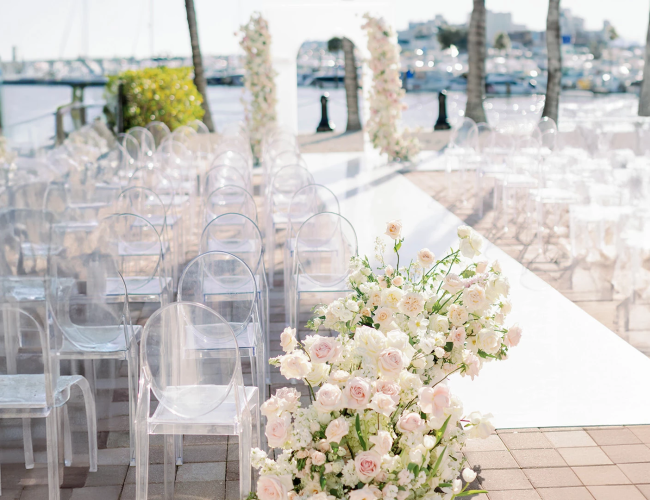 Elegant outdoor wedding setup by the harbor with clear acrylic chairs and a white aisle lined with pale pink roses and greenery.