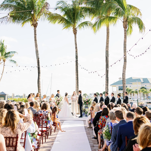A wedding ceremony outdoors by palm trees, guests seated on both sides, aisle runner, string lights, sunny day near a marina or resort.