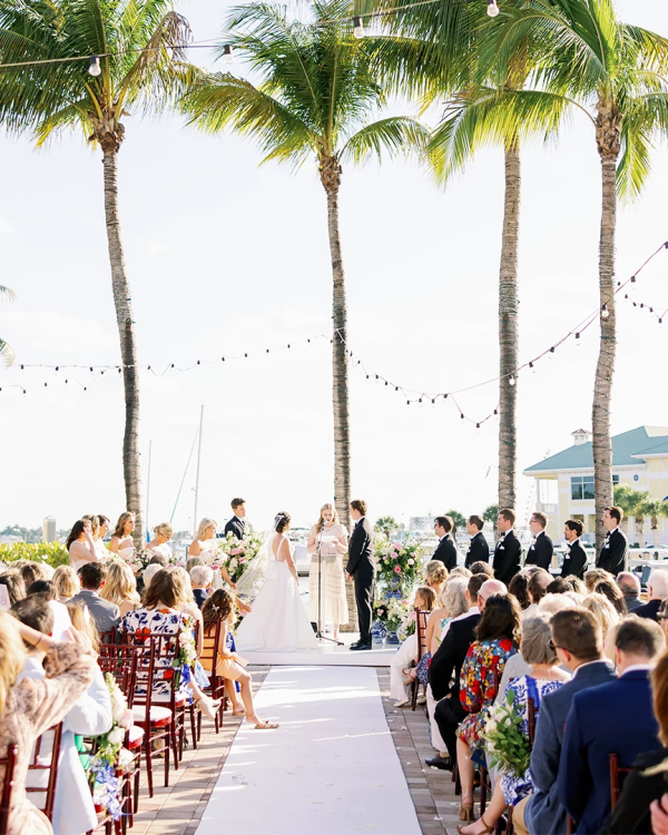 A wedding ceremony outdoors by palm trees, guests seated on both sides, aisle runner, string lights, sunny day near a marina or resort.