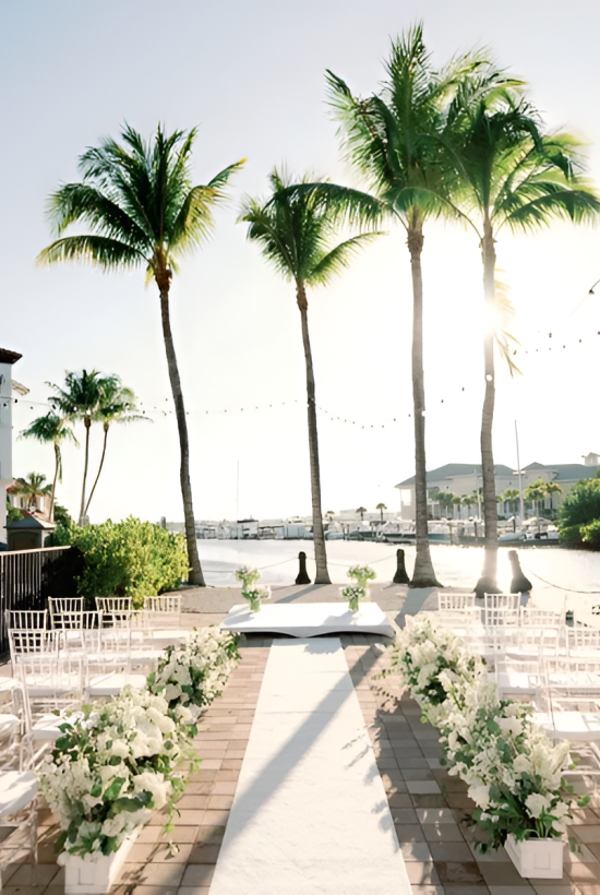 A tropical outdoor wedding setup with white chairs and floral aisles along a wooden pathway, palm trees, string lights, and a sunny waterfront backdrop.