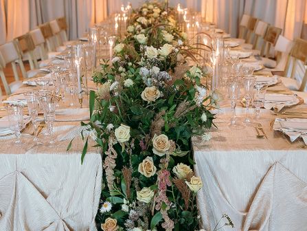 A long banquet table adorned with a lush floral centerpiece of cream and white roses, greenery, and string lights, set for a formal event.