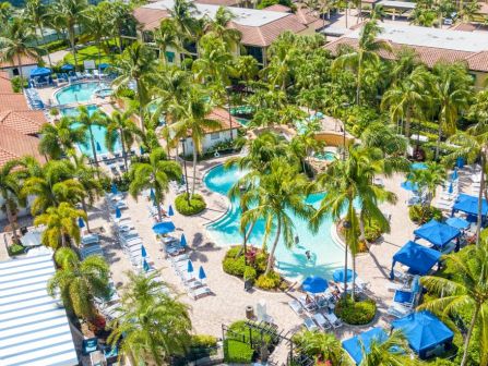 A tropical resort with numerous palm trees, pools shaped in waves, sun loungers under blue umbrellas, and lush greenery surrounding sunlit buildings. end.