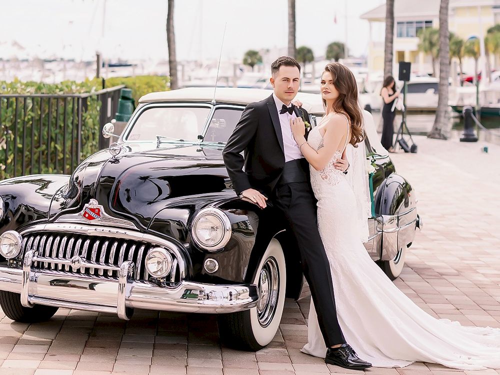 A newlywed couple posing by a vintage black car, man in a suit and bow tie, woman in a white wedding dress, waterfront background.