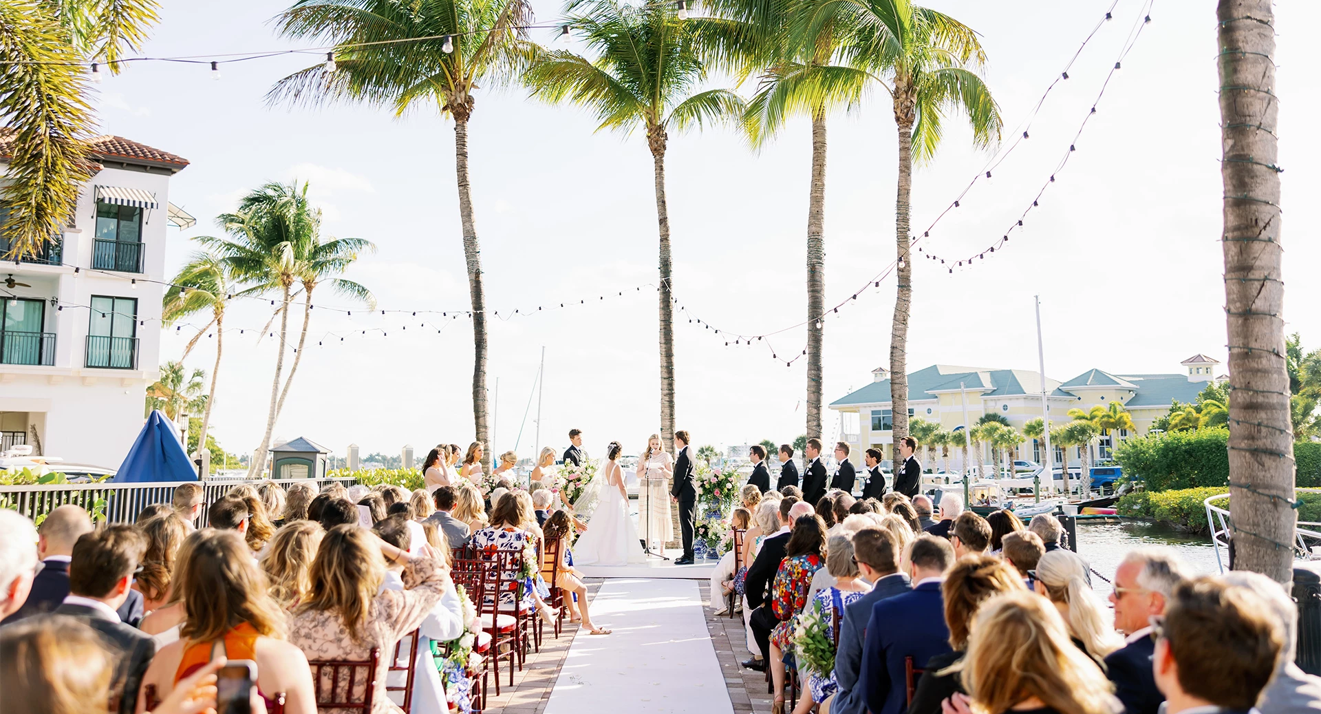 A beachside wedding ceremony with guests seated along a white aisle, tropical palm trees, string lights, and a bright seaside backdrop.
