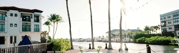 A sunny coastal wedding setup with white chairs, floral aisle decor, palm trees, and string lights by the water.