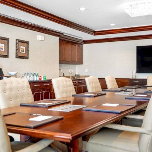 A modern conference room with a long wooden table, beige upholstered chairs, a wall-mounted TV, and coffee service in the back&mdash;bright and professional.