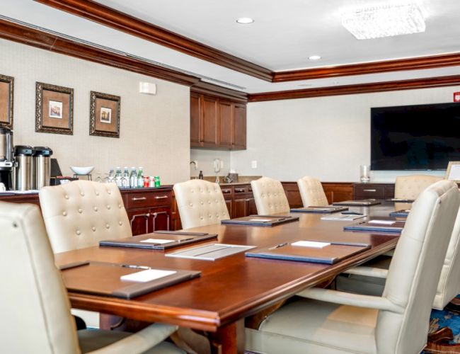 A modern conference room with a long wooden table, beige upholstered chairs, a wall-mounted TV, and coffee service in the back&mdash;bright and professional.