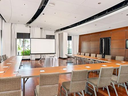 A modern conference room with a large U-shaped table, many chairs, a projector screen, and wooden accents.
