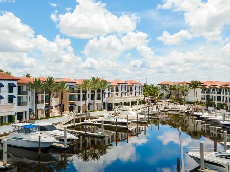 A marina with boats docked along calm, mirror-like water, surrounded by low-rise buildings and palm trees under a bright, cloudy sky.