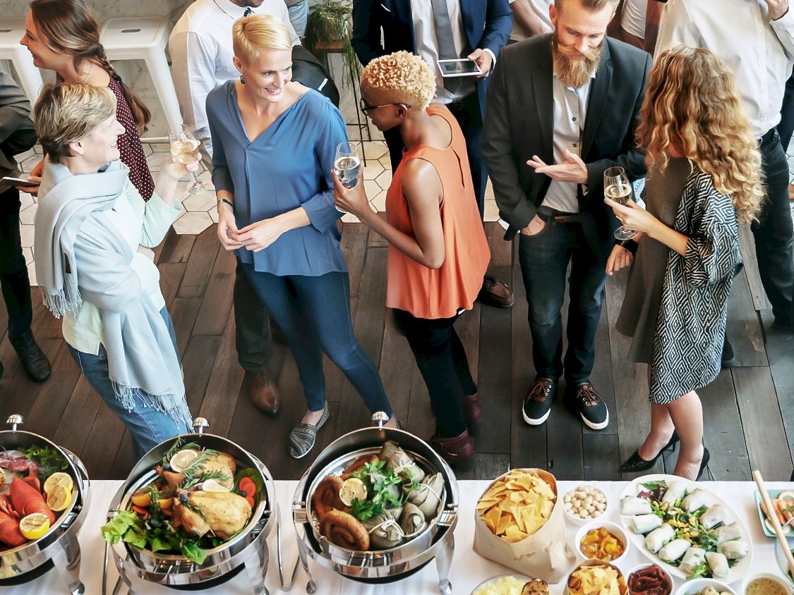 A group of well-dressed people mingling at a catered buffet with bowls, salads, and finger foods on display.