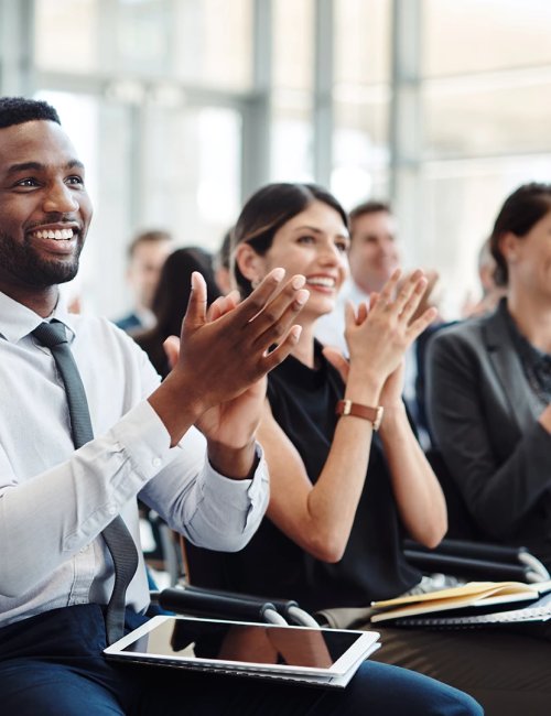 People smiling and clapping in a modern conference room, seated with notebooks and tablets, enjoying a presentation or speech.