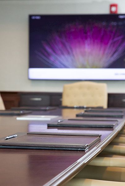 A conference room with a long table, beige chairs, and multiple tablets or notebooks laid out; a large screen is mounted on the wall.