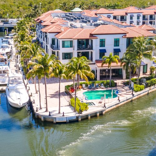Aerial view of a sunny marina with canals, luxury white buildings with red-tile roofs, palm trees, yachts docked along the water, and a central pool area. End.
