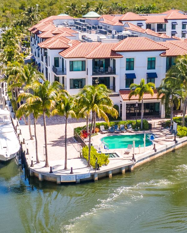 Aerial view of a sunny marina with canals, luxury white buildings with red-tile roofs, palm trees, yachts docked along the water, and a central pool area. End.