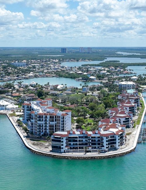 A curved island with multi-story buildings sits in turquoise water, connected by canals, with boats gliding around and a bright, cloud-filled sky.