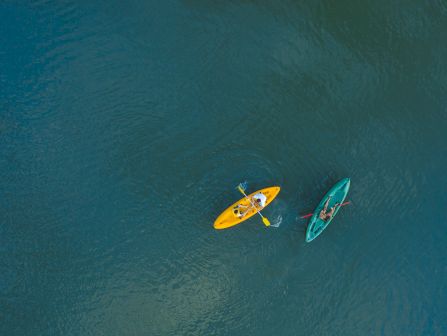 Two kayakers paddle on a clear blue-green ocean from above, one in a yellow kayak and one in teal.
