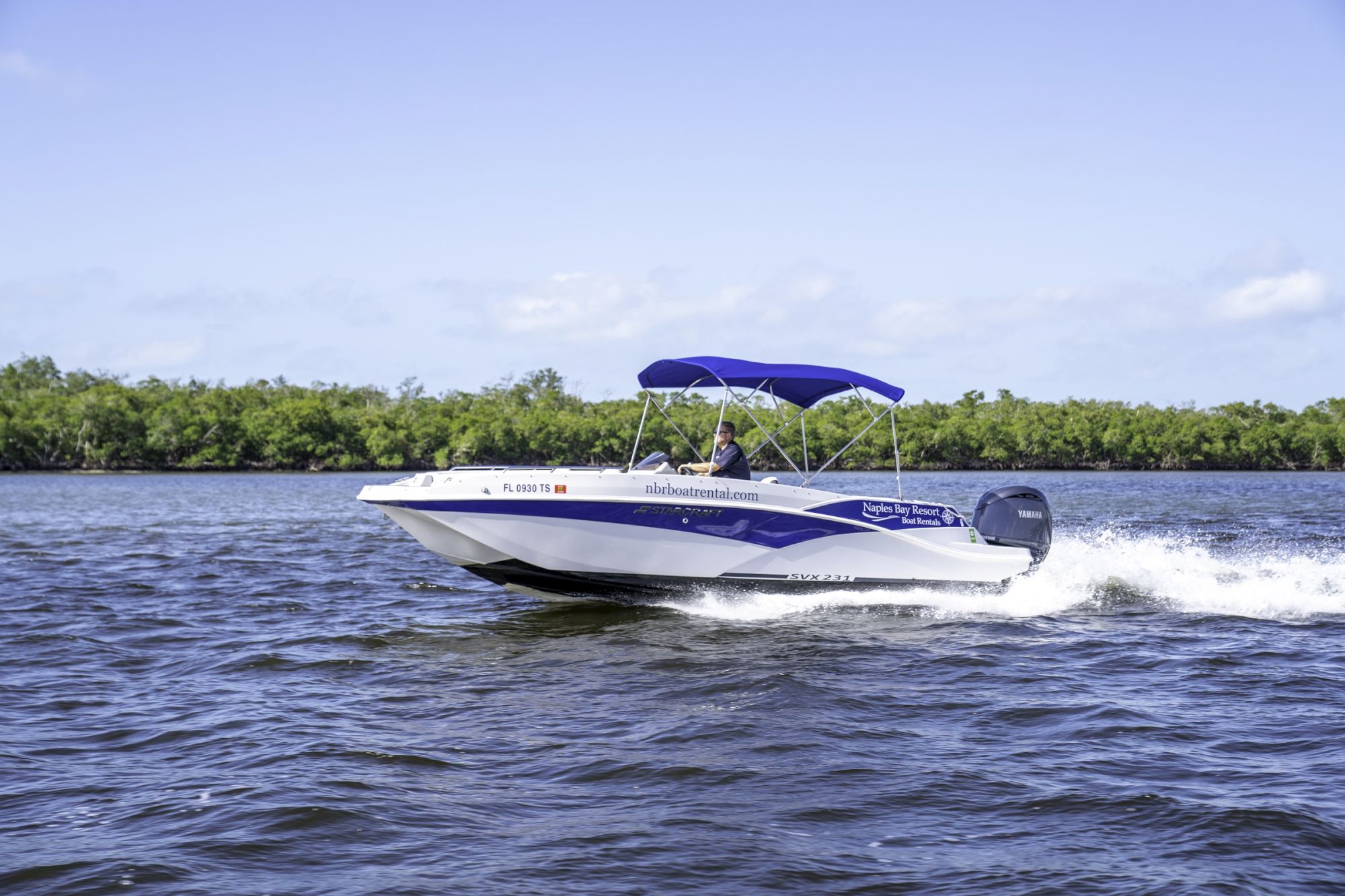 A speedboat with a blue canopy cruises fast on dark water, leaving a white wake near a distant shoreline of low mangroves.