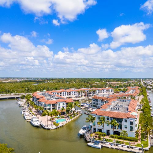 An aerial view of a coastal canal town with colorful multi-story buildings along the water, boats docked at the piers, and a bright, sunny sky.
