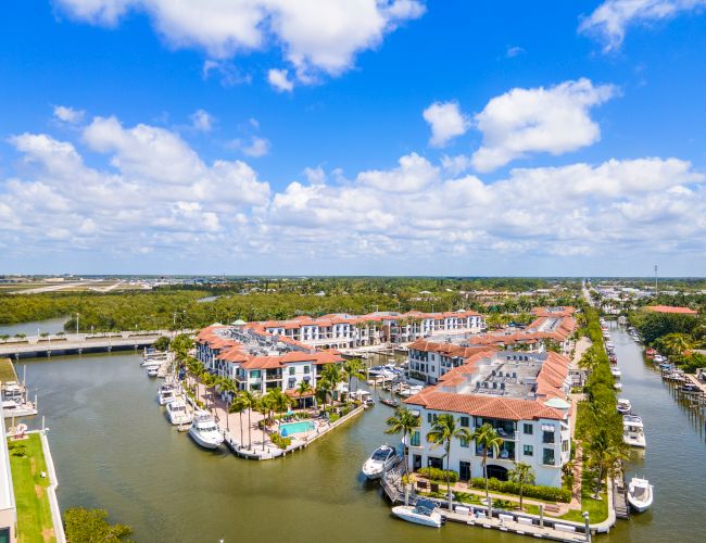 An aerial view of a coastal canal town with colorful multi-story buildings along the water, boats docked at the piers, and a bright, sunny sky.