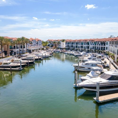 A tranquil marina with several boats docked along calm water, flanked by white Mediterranean-style townhomes with red-tiled roofs.
