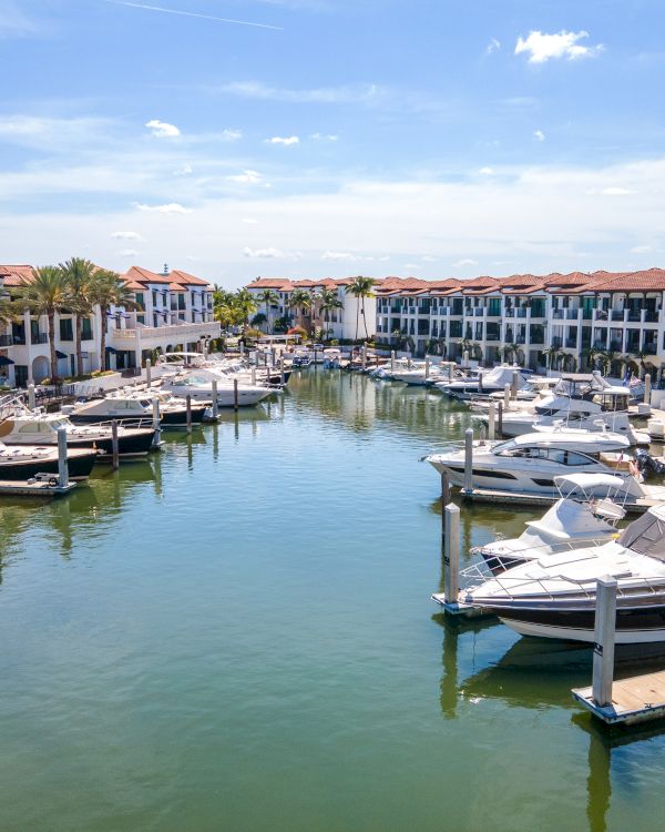 A tranquil marina with several boats docked along calm water, flanked by white Mediterranean-style townhomes with red-tiled roofs.