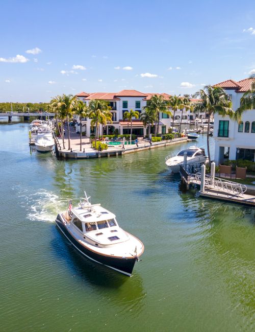 A sunny canal scene with boats cruising past waterfront houses, palm trees lining the shore, and calm greenish water reflecting a bright sky.