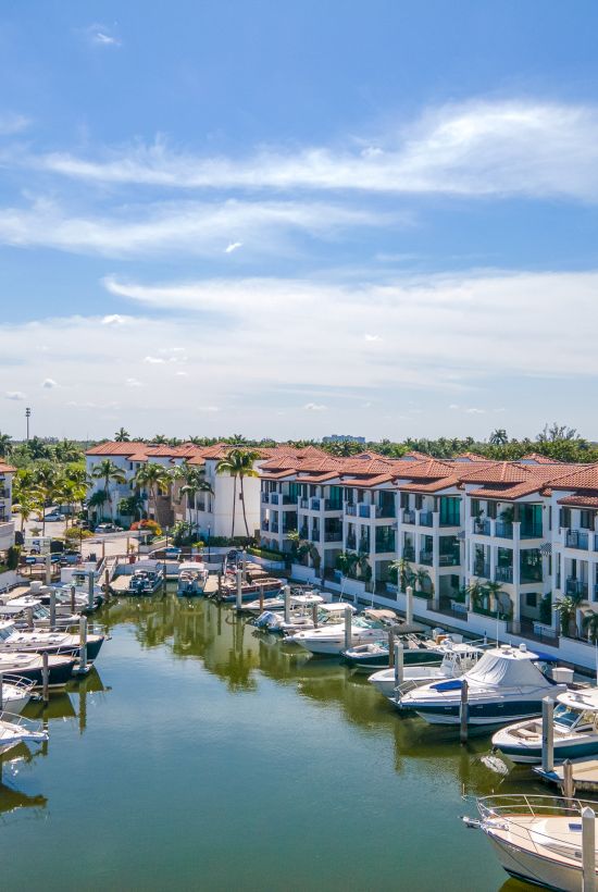 A sunny marina surrounded by three-story pastel buildings, boats docked along calm water, and a clear blue sky above.