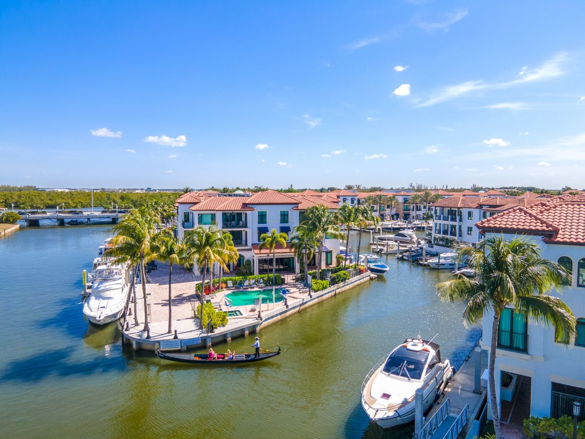 A sunny coastal marina with luxury homes, palm trees, and several yachts docked along calm waters.