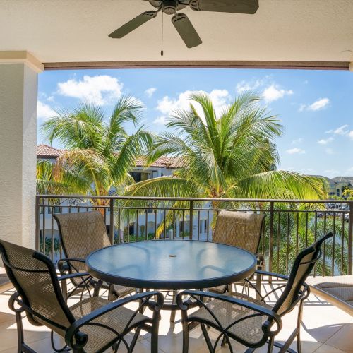 A covered balcony with a round glass table and four metal chairs, overlooking palm trees and a sunny poolside view.