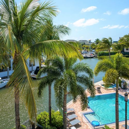 A sunny resort scene with palm trees, a pool, and a canal surrounded by buildings and boats.