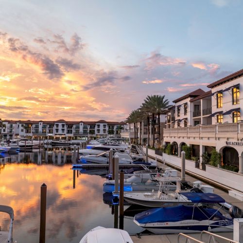 Sunset over a marina with calm water, boats docked along piers, and elegant white buildings lining the waterfront.