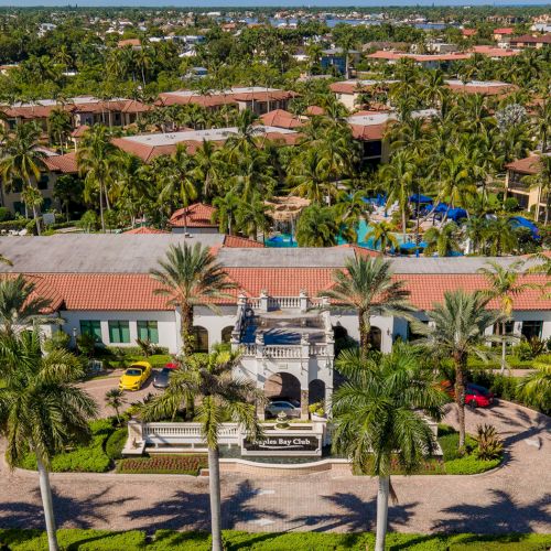 A tropical resort with red-tiled roofs, palm trees, a central entrance, and a pool area surrounded by manicured lawns and sunny grounds.