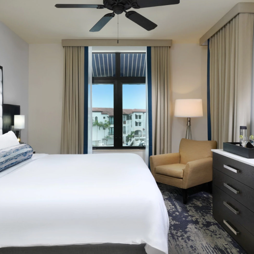 A modern hotel room with a white bed, bedside lamps, a ceiling fan, a window with curtains, a beige chair, and a dark dresser with a wall-mounted TV.