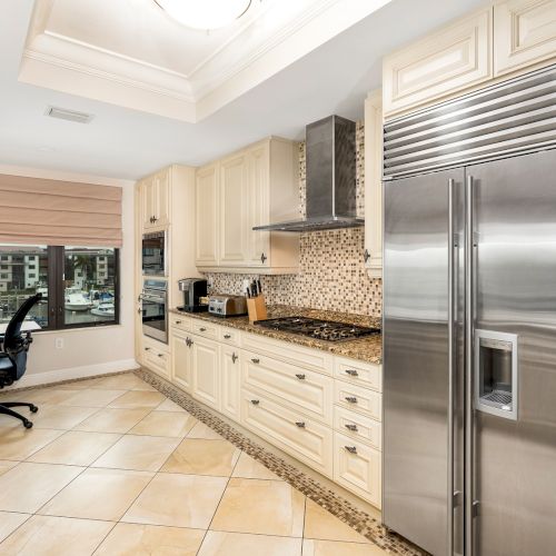A modern kitchen with light wood cabinets, granite countertops, tile floor, stainless steel fridge, and a small work desk by a window.