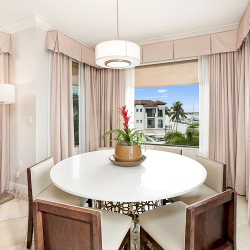 A bright dining nook with a round white table, four cushioned chairs, pink drapes, a pendant light, and a view of a sunny outdoor scene through a window.