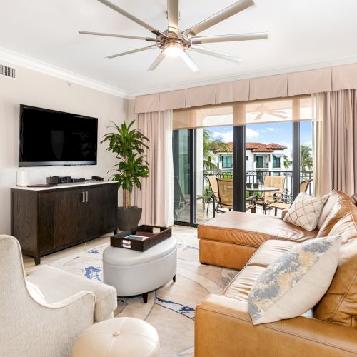 A bright living room with a tan leather sectional, beige armchairs, a wall-mounted TV, a dark wood console, a ceiling fan, and a sliding glass door to a balcony. End.