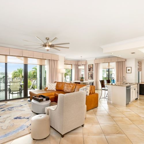 Spacious, sunlit open-plan living room with beige tiled floor, tan sofa, gray armchairs, a kitchen island, ceiling fan, and glass doors to a balcony overlooking greenery.