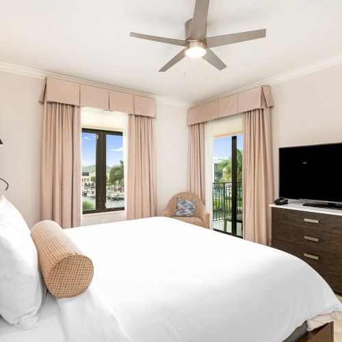 A bright hotel room with a king-size bed, beige headboard, white linens, pink curtains, ceiling fan, and a dresser with a flat-screen TV, plus a balcony view.