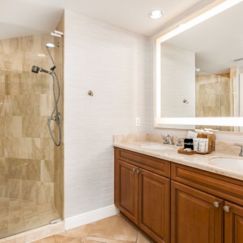 A modern bathroom with a glass-enclosed shower, beige tiled walls and floor, a wooden double-sink vanity, and a large backlit mirror above the counter.